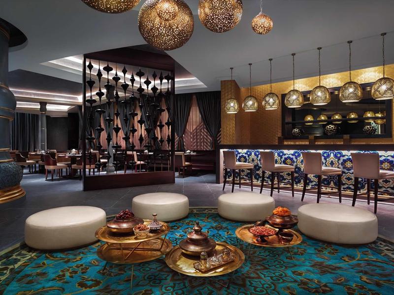 Moderner Loungebereich mit rundem Teppich, weißen Hockern, goldenem Kerzenhaltern und Bar im Hintergrund.