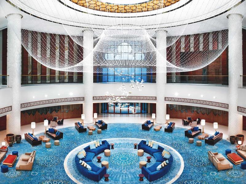 Großer runder Raum mit blauer Teppichfläche, weißen Säulen und einem glänzenden Glasdach.