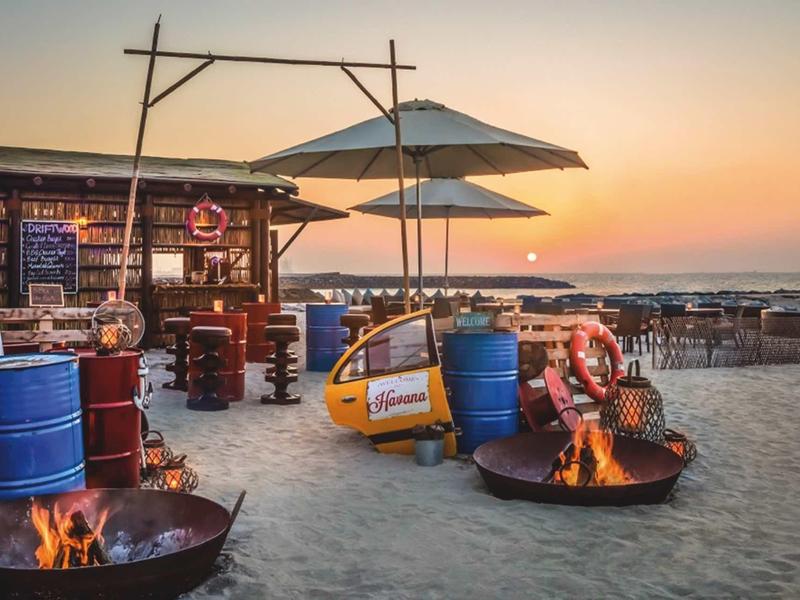Strandbar mit Holzbuden, Feuerstellen, bunten Fässern und Sonnenuntergang am Meer.
