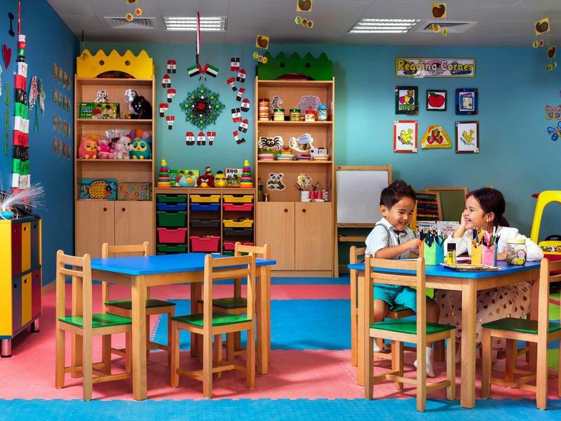 Bunter Kindergartenraum mit kleinen Tischen, Stühlen, Spielzeug und zwei Kindern beim Spielen.