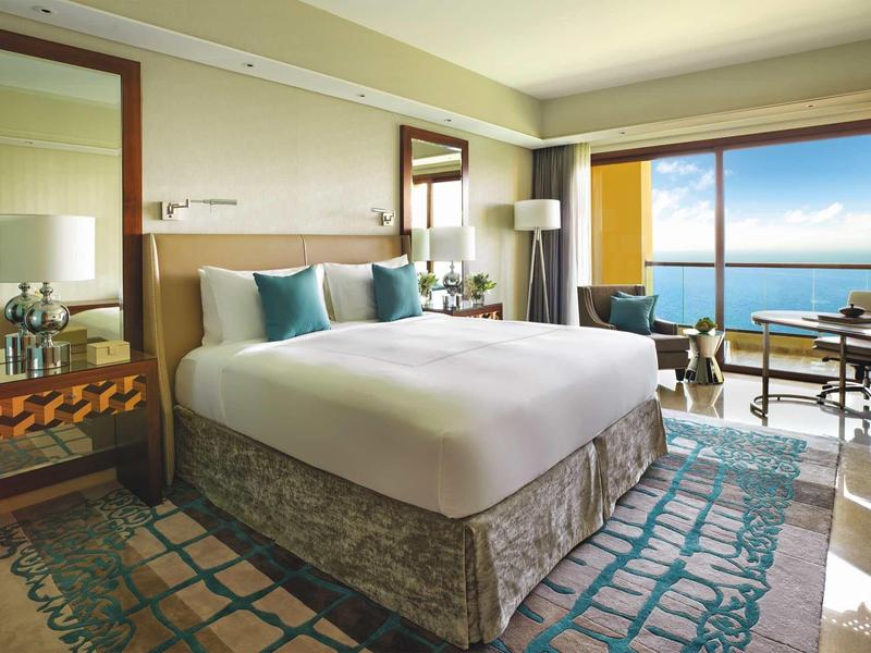 Hell eingerichtetes Schlafzimmer mit großem Bett, Teppich und Meerblick durch bodentiefe Fenster.