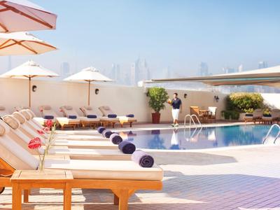 Hotel pool with lounge chairs, umbrellas, and a city skyline under a clear sky.