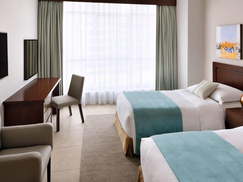 Jasny pokój hotelowy z dwoma łóżkami, biurkiem i fotelem przy dużym oknie.