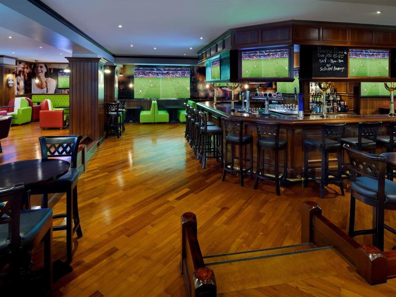 Nowoczesny bar hotelowy z drewnianą podłogą, miejscami do siedzenia i kilkoma telewizorami.