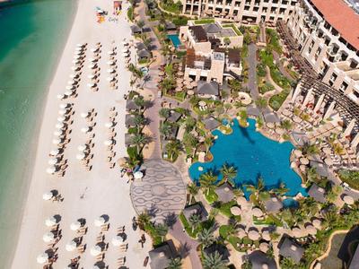 Strand mit weißen Liegen, angrenzender großer Pool und Hotelanlage mit grünen Pflanzen und Wegen.