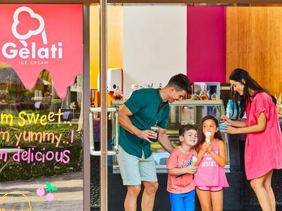Familie mit zwei Kindern steht lächelnd vor Eisladen mit bunten Wänden und Schriftzug "Gelati".