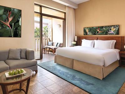 Helles Schlafzimmer mit grauem Sofa, großem Bett, blauem Teppich und Pflanzenbildern an der Wand.