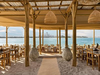 Otwarta restauracja na plaży z drewnianymi meblami z widokiem na morze i panoramę miasta.