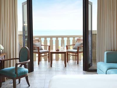 Hotelzimmer mit blauem Sessel, Holztischen und offener Tür zum Balkon mit Meerblick.