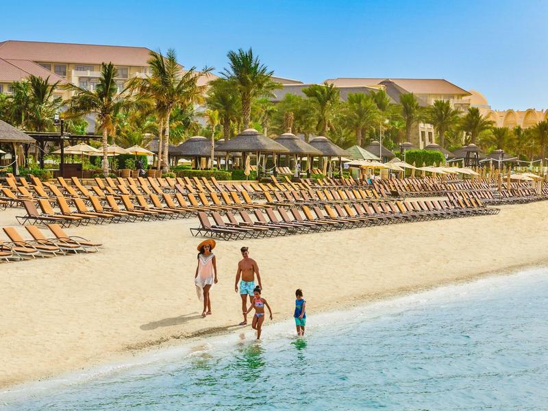Strand mit Sand, Palmen, Liegestühlen und drei Personen im seichten Wasser an einem sonnigen Tag.