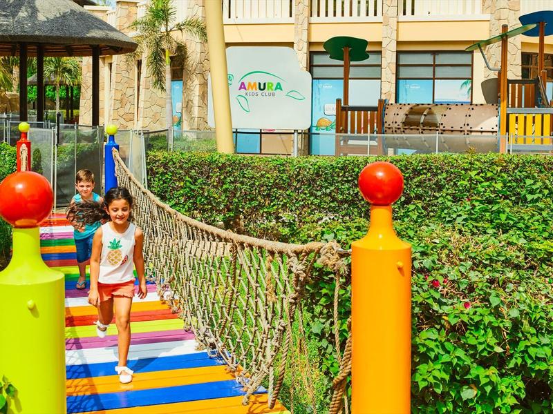 Kinder überqueren eine bunte Hängebrücke in einem Spielplatz mit grünem Busch und Holzspielgerät.