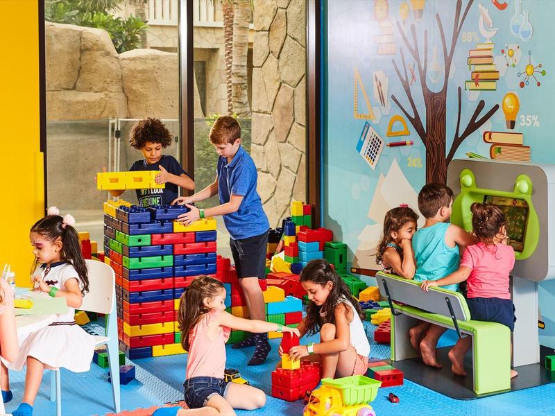 Kinder spielen mit bunten Bausteinen und sitzen an Tischen in einem hellen, farbenfrohen Raum.