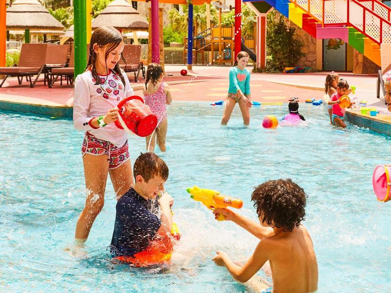Kinder spielen im flachen Schwimmbecken mit Wasserpistolen und Schaufeln an einem sonnigen Tag.