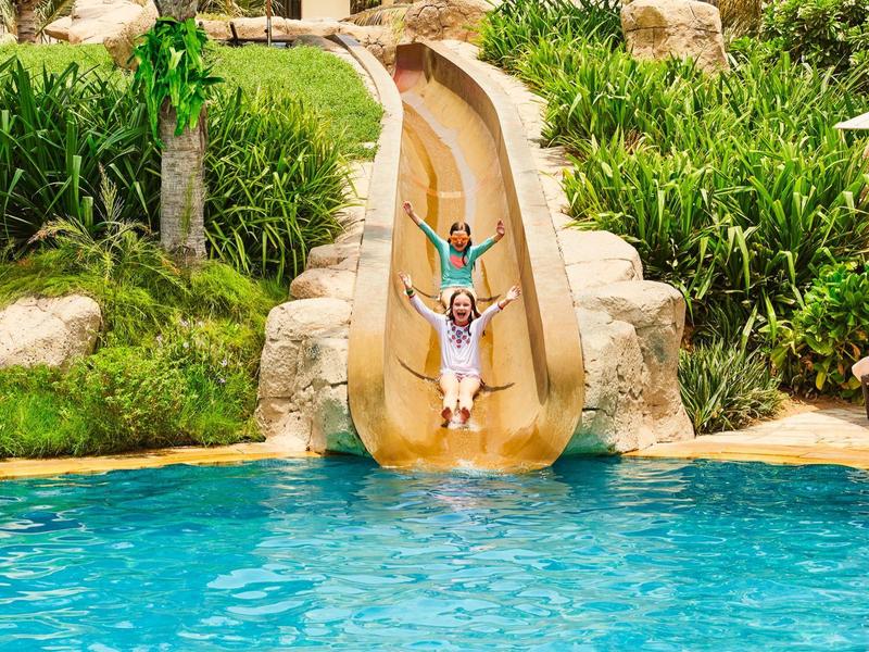 Zwei Kinder rutschen eine braune Wasserrutsche hinab in einen blauen Pool, umgeben von grüner Vegetation.
