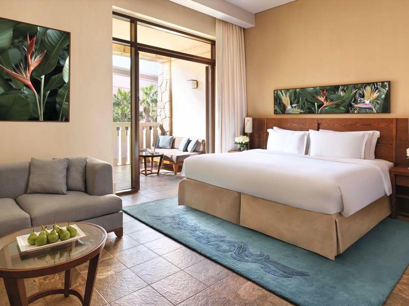 Helles Schlafzimmer mit grauem Sofa, großem Bett, blauem Teppich und Pflanzenbildern an der Wand.