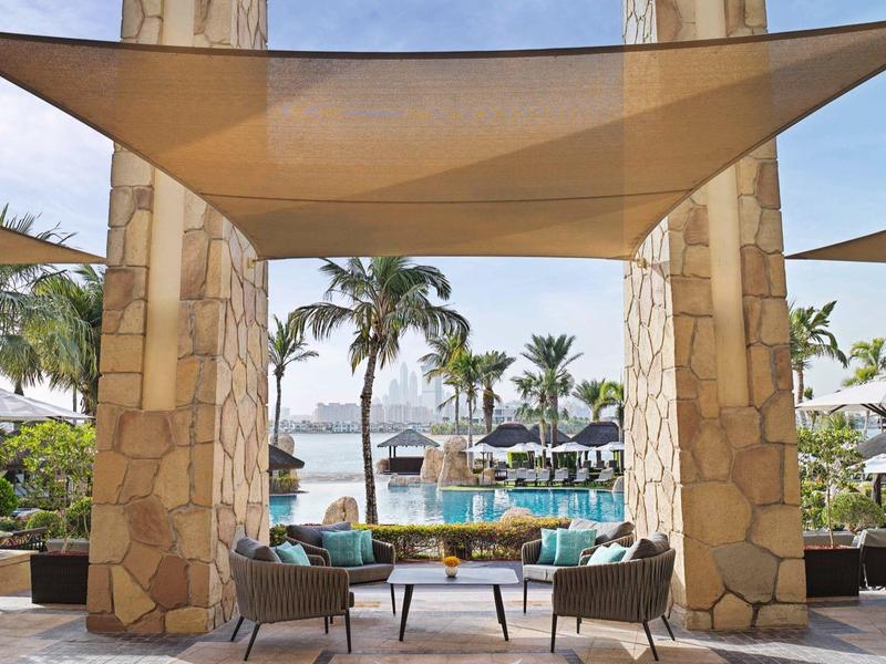 Große Steinsäulen mit Sonnensegel, Lounge-Möbel und Blick auf Palmen und Meer im Hintergrund.