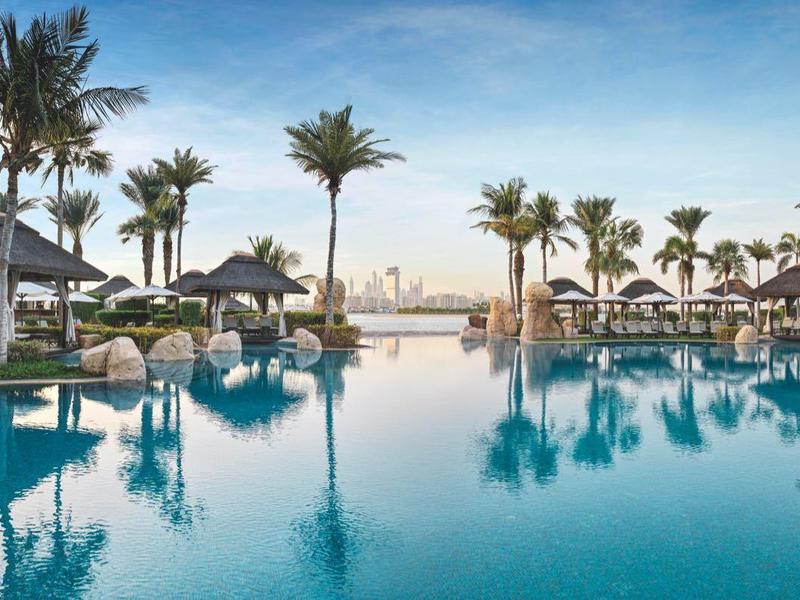 Großer Pool mit hellblauem Wasser, Palmen und weißen Sonnenschirmen unter blauem Himmel.