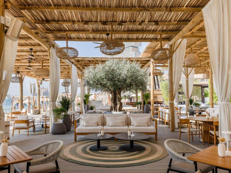 Helles, offenes Restaurant mit Holzdecke, weißen Vorhängen, Pflanzen und zentralem Olivenbaum.