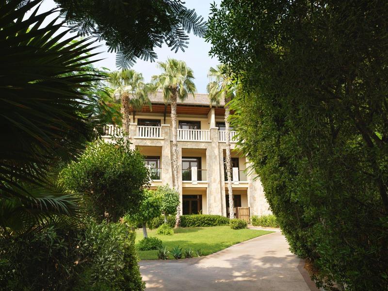 Mehrstöckiges Gebäude mit Balkon, umgeben von grünen Bäumen und einem Gehweg im Vordergrund.