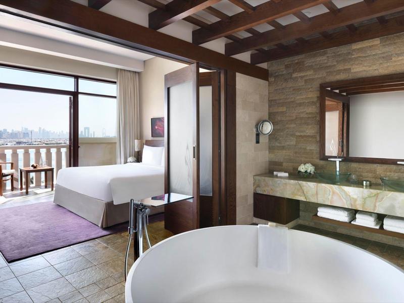 Modernes Schlafzimmer mit Balkon, großem Bett, freistehender Badewanne und Natursteinwand im Bad.