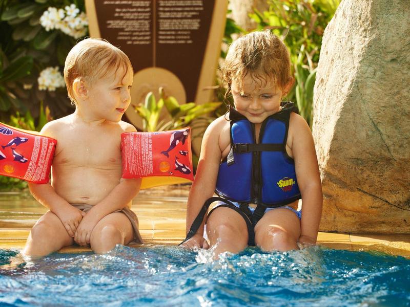 Zwei Kinder mit Schwimmflügeln sitzen am Poolrand und plantschen im Wasser.