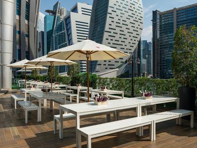 Terrazza con tavoli bianchi, panche e ombrelloni davanti a grattacieli moderni.