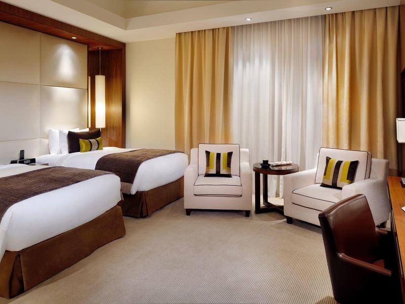 Nowoczesny pokój hotelowy z dwoma łóżkami pojedynczymi, fotelami i biurkiem przy ciepłym świetle.