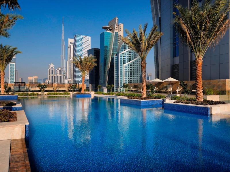 Duży basen z palmami na tle nowoczesnej miejskiej panoramy przy czystym niebie.