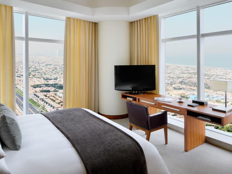 Pokój hotelowy z dużym łóżkiem, biurkiem, telewizorem i panoramicznymi oknami z widokiem na miasto.