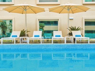 Čtyři bílé lehátka s modrými polštáři pod slunečníky u bazénu před hotelem.