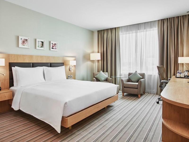 Modern hotel room with large double bed, two armchairs, and desk by a wide window.