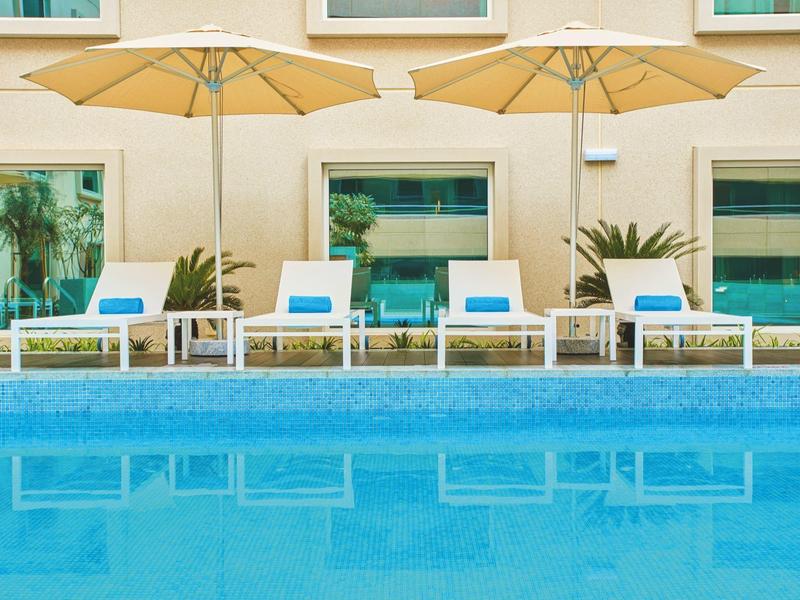 View of a pool with lounge chairs and umbrellas in front of a hotel building.