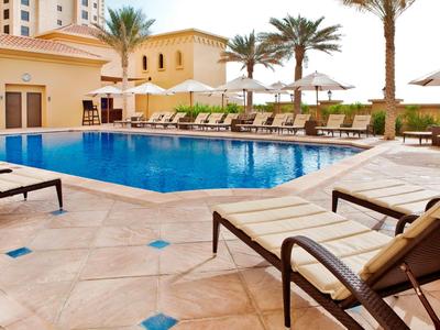 Moderner Außenpool mit Liegestühlen, Sonnenschirmen und Palmen in einem Hotelkomplex.