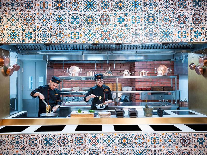Deux chefs dans une cuisine moderne avec des carreaux colorés et des étagères en bois.