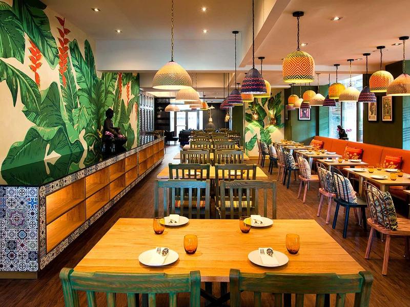 Restaurant moderne avec décoration murale tropicale, chaises en bois et colorées autour de la table.