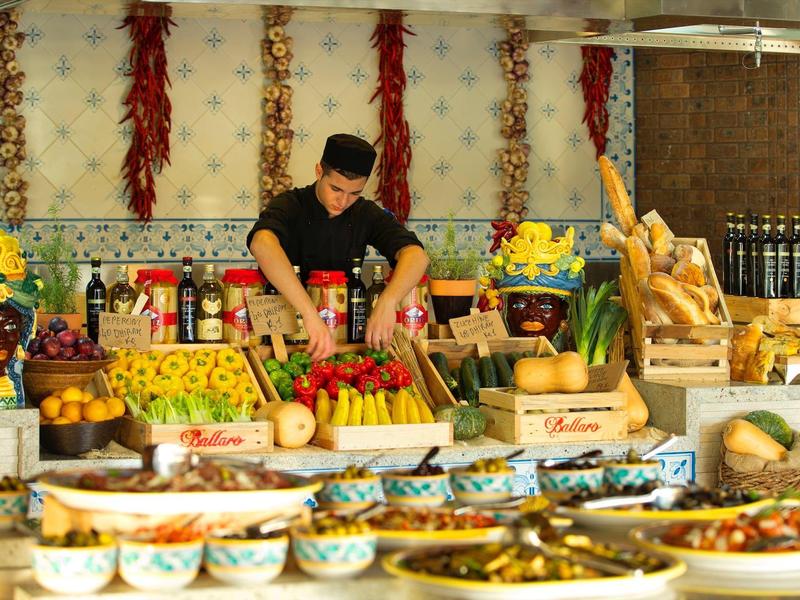 Marktstand mit bunter Gemüse- und Obstauslage, Gewürzen und Brotsorten, Verkäufer mischt Zutaten.