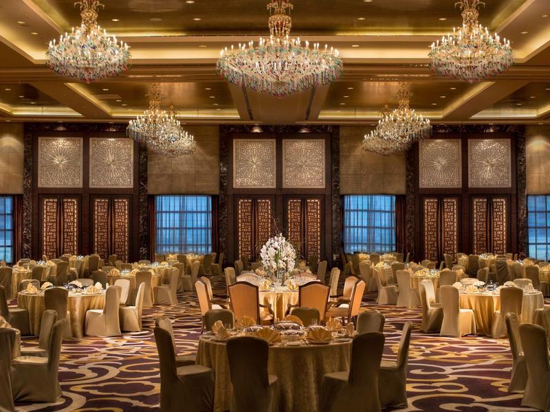 Großer festlicher Saal mit mehreren runden Tischen, goldenen Stühlen und Kronleuchtern an der Decke.