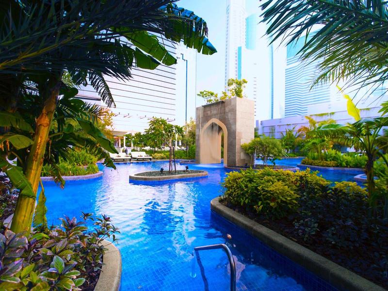 Blauer Pool mit Palmen, grünen Sträuchern und modernen Hochhäusern im Hintergrund bei Tageslicht.