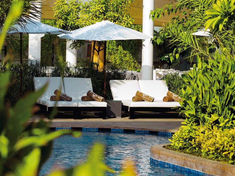 Einladende Poollandschaft mit weißen Sonnenschirmen und gemütlichen Sitzmöbeln, umgeben von grünem Pflanzen.