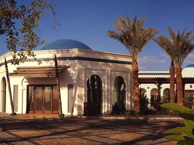 A white building with a round roof, surrounded by palm trees and plants in sunlight.