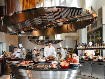 Moderní hotelová kuchyně s kuchaři připravujícími a prezentujícími pokrmy na centrálním kuchyňském ostrově.