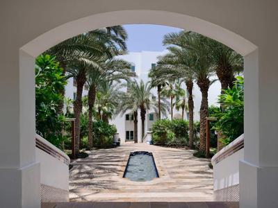 Pohled skrz oblouk na promenádu s palmami a vodním prvkem v hotelovém areálu.