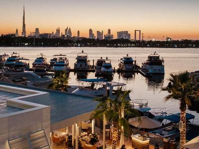 Pohled na osvětlenou hotelovou terasu u vody s loděmi a panoramatem města při západu slunce.