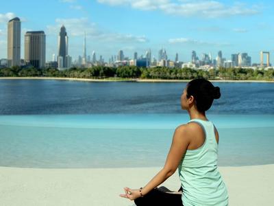 Žena v jóga pozici u břehu s výhledem na moderní panorama města přes vodu.