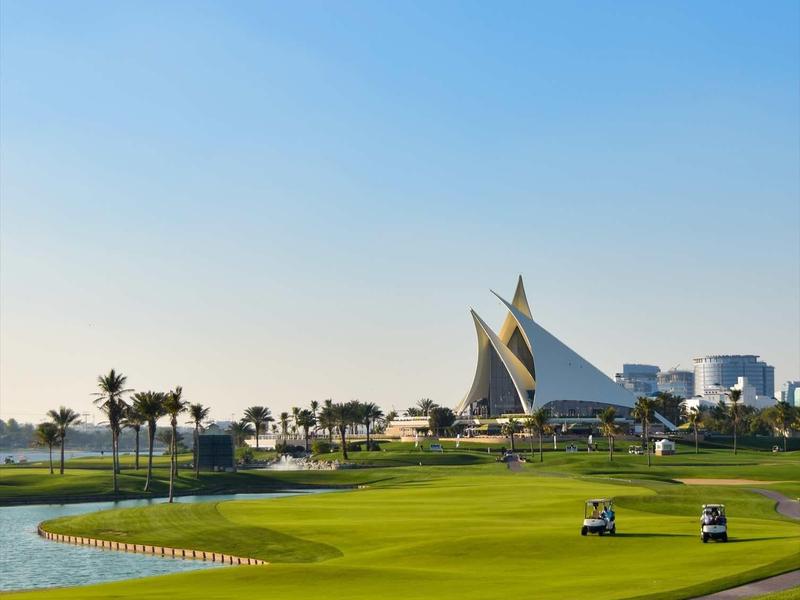 Zelené golfové hřiště s dvěma golfovými vozíky a výraznou moderní architekturou v pozadí pod jasnou oblohou.