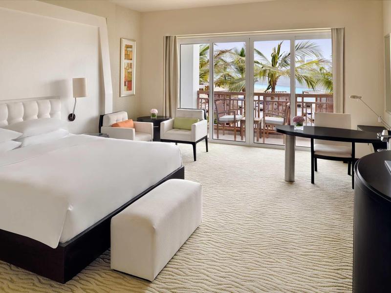 Moderní hotelový pokoj s velkou postelí, posezením a balkonem s výhledem na palmy.
