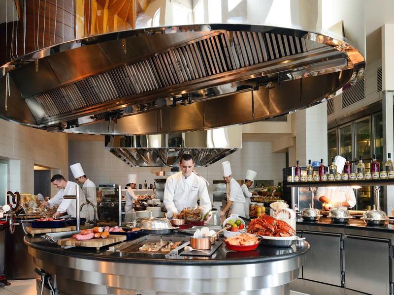 Moderní hotelová kuchyně s kuchaři připravujícími a prezentujícími pokrmy na centrálním kuchyňském ostrově.