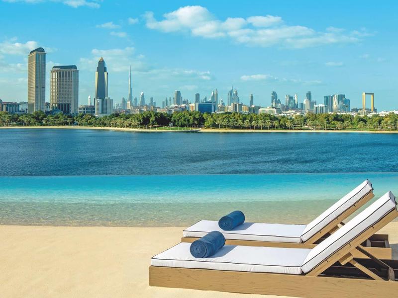 Dvě lehátka s modrými polštáři na písečné pláži s výhledem na městskou oblohu a modrou oblohu.