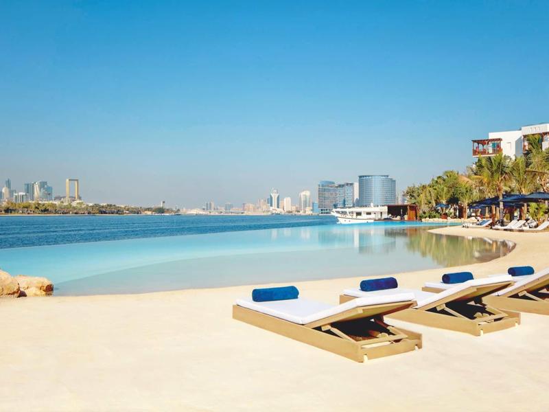 Luxusní lehátka na klidné pláži s výhledem na moderní město a průzračnou modrou vodu.