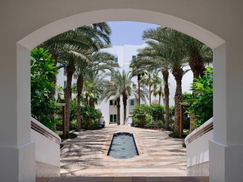 Pohled skrz oblouk na promenádu s palmami a vodním prvkem v hotelovém areálu.